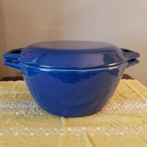 May include: A cobalt blue, lidded Dutch oven. The ceramic pot has two handles and sits on a yellow tablecloth with white floral embroidery. The background is a neutral tan color.
