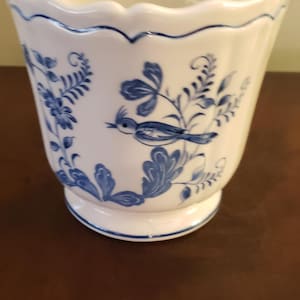 May include: White ceramic planter with a scalloped rim and a blue floral and bird design. The planter has a flared shape and a pedestal base. A decorative piece for home decor.