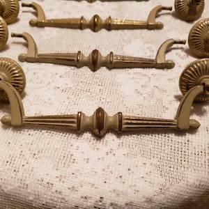 May include: A collection of antique cabinet hardware, including drawer pulls and matching knobs. The hardware features a cream-colored finish with gold accents. The drawer pulls have a decorative, ornate design, and the knobs are round with a fluted pattern.
