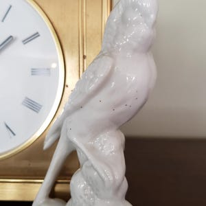 May include: A white ceramic figurine of a bird perched on a textured base. The bird has detailed feathering and is set against a gold-framed clock.