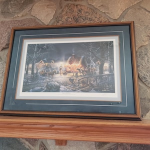 May include: Framed print depicting a winter scene with a warmly lit cabin, horses, and figures in a snowy landscape. The artwork is set within a dark green mat and a brown wooden frame, displayed on a wooden shelf against a stone wall.