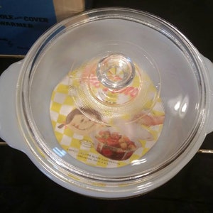 K&ouml;nnte beinhalten: Eine wei&szlig;e Auflaufform mit einem Glasdeckel und goldenen Griffen. Der Deckel hat ein gelbes und wei&szlig;es Karomuster mit einem Comic-M&auml;dchen darauf. Die Form ist mit "Fire King White 1 1/2 Qt Casserole - Cover Candle Warmer" beschriftet.