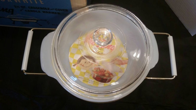 K&ouml;nnte beinhalten: Eine wei&szlig;e Auflaufform mit einem Glasdeckel und goldenen Griffen. Der Deckel hat ein gelbes und wei&szlig;es Karomuster mit einem Comic-M&auml;dchen darauf. Die Form ist mit "Fire King White 1 1/2 Qt Casserole - Cover Candle Warmer" beschriftet.