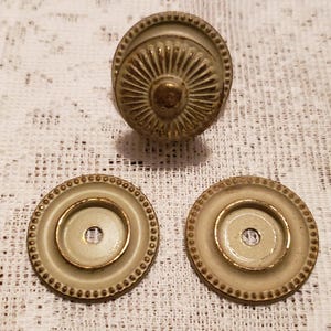 May include: Antique brass-toned cabinet hardware set, including a decorative knob and two backplates. The knob features a detailed, sunburst-like design. The backplates have a circular shape with a beaded edge and a central hole.