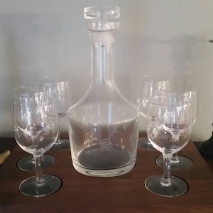 May include: A set of six clear glass wine glasses and a clear glass decanter with a black stopper on a wooden tray. There are two black metal decorative items on the left and a black metal candle holder on the right.