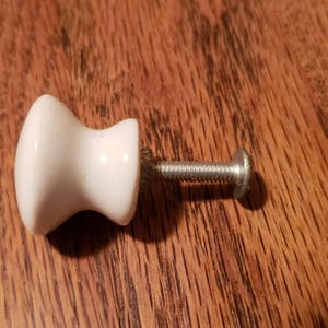 May include: A white ceramic cabinet knob with a decorative shape, attached to a silver-colored screw. The knob is set against a wooden surface.