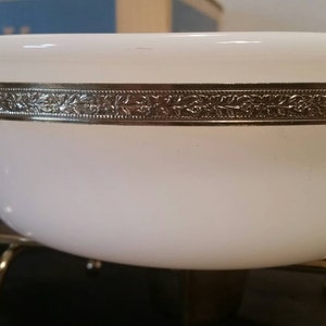 K&ouml;nnte beinhalten: Wei&szlig;e Keramiksch&uuml;ssel mit goldfarbenem Rand und Blumenmuster. Die Sch&uuml;ssel steht auf einem goldfarbenen Metallst&auml;nder mit drei Beinen.