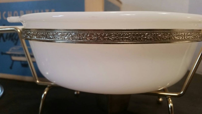 K&ouml;nnte beinhalten: Wei&szlig;e Keramiksch&uuml;ssel mit goldfarbenem Rand und Blumenmuster. Die Sch&uuml;ssel steht auf einem goldfarbenen Metallst&auml;nder mit drei Beinen.