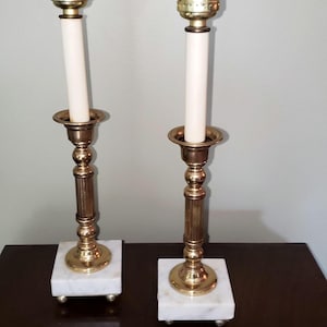 May include: A pair of brass candlestick lamps featuring a tall, slender central column with a fluted design, mounted on a square white marble base with small spherical feet. Each lamp includes a gold-toned metal socket and a white candle sleeve.