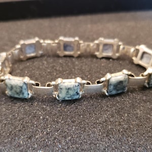 May include: A silver bracelet featuring square, speckled blue-green stones. The bracelet is composed of rectangular links connecting the square stones, creating a flexible design. The stones have a mottled appearance, adding visual interest to the jewelry.