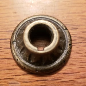May include: A vintage brass-toned furniture knob with a circular design. The knob features a central opening and a decorative, ridged pattern around the base. The knob is likely for a drawer or cabinet.