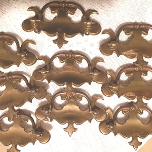May include: A collection of eight antique brass drawer pulls. Each pull features a decorative, ornate design with a curved handle and a detailed backplate. The pulls are a warm, golden color, suggesting age and use.