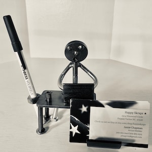 Puede incluir: Una escultura de metal de una persona sentada en un escritorio con una computadora portátil, un portaplumas y un porta tarjetas de visita. El porta tarjetas de visita tiene una tarjeta de visita con el texto "Happy Skraps" y la información de contacto del artista.