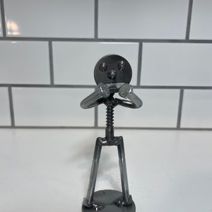 May include: A metal sculpture of a person with a round head and arms crossed over their chest. The sculpture is made of nuts, bolts, and screws.