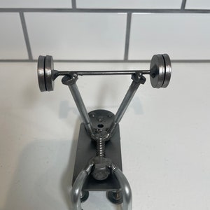 Peut inclure: Une sculpture métallique d'un haltérophile, fabriquée à partir de diverses pièces métalliques. La figurine est représentée en position de levage, avec une barre chargée de deux disques. La sculpture est en métal argenté et repose sur une surface blanche.