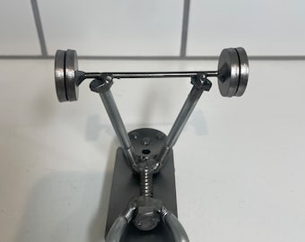 May include: A metal sculpture of a weightlifter, crafted from various metal pieces. The figure is depicted in a lifting pose, with a barbell loaded with two weight plates. The sculpture is made of silver-colored metal and sits on a white surface.
