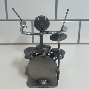 Pode incluir: Escultura de metal artesanal de um baterista tocando bateria. A escultura é feita de várias peças de metal, incluindo parafusos e arruelas. A figura do baterista tem uma cabeça redonda, braços e pernas e está segurando baquetas. A bateria inclui um bumbo, uma caixa e pratos.