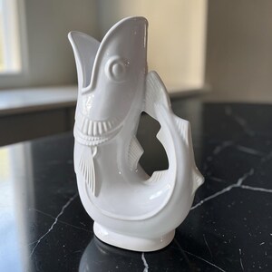 May include: A glossy white ceramic pitcher shaped like a fish. The vessel features a wide, open mouth at the top serving as the spout, with molded scales, fins, and a circular eye detail. The handle is formed by the curved tail section of the fish.