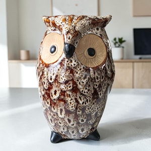 May include: A ceramic owl figurine featuring a glossy, mottled brown and cream glaze. The owl has large, circular tan eyes with black pupils, a pointed black beak, and two small black feet at the base. The textured surface mimics feathers.