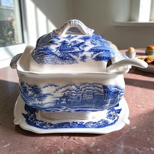 May include: A cream-colored ceramic tureen with a matching lid, ladle, and rectangular base plate. The set features a blue transferware pattern depicting a castle landscape, trees, and a boat. The tureen has scalloped edges and side handles.