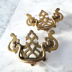 May include: Two antique brass drawer pulls with ornate, openwork designs. Each pull features a curved handle and decorative flourishes, showcasing a vintage aesthetic. The hardware is a warm, golden color, suggesting age and use.