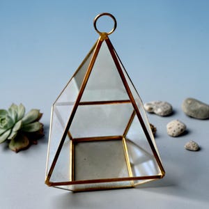 May include: A geometric terrarium container featuring clear glass panels framed in thin gold-toned metal. The structure has a pyramid-like shape with a square base and a small circular ring at the top for hanging or carrying purposes.