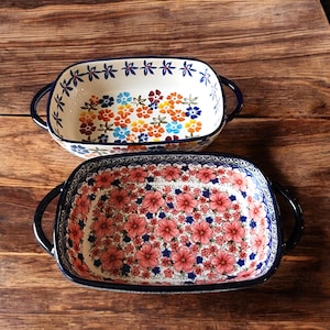 May include: Two rectangular ceramic baking dishes with dark blue handles and rims. One dish features a floral pattern with orange, blue, and yellow flowers. The other has a pattern of pink flowers with blue accents. Both are on a wooden surface.