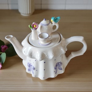 May include: A white ceramic teapot with a ruffled skirt design, adorned with purple bows and polka dots. The lid features a miniature teapot, teacup, and a bird figurine. A small vase with flowers is also on the lid.