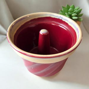 May include: A deep red ceramic bundt pan with a glossy finish and a central chimney. The rim features a contrasting cream-colored glaze that extends slightly down the exterior. The exterior surface displays a subtle spiral or fluted texture.