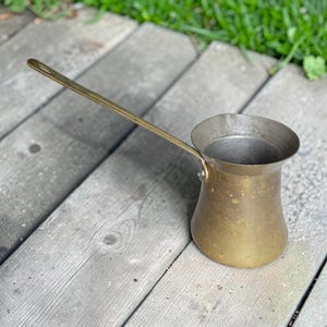 May include: A brass coffee pot with a long, slender handle and a wide, flared rim. The body of the pot has a tapered, cylindrical shape with a metallic, weathered finish. The handle is attached to the side of the pot with two visible rivets.