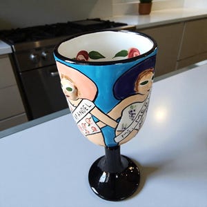 May include: A hand-painted ceramic goblet with a black base and a wide, detailed bowl. The bowl features two figures in hats, holding hands, with the words "MF ANDEL" and "MS. MER" on their clothing. The goblet is decorated with blue, white, and flesh tones.