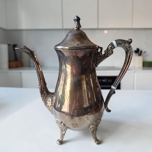 May include: A silver-toned metal coffee pot featuring an ornate, curved handle with two white accents, a long slender spout, and a pointed lid. The body of the pot is smooth and reflective, supported by three decorative, detailed feet at the base.