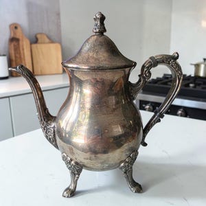 May include: A silver-plated teapot with a polished, reflective surface, featuring an ornate handle, a curved spout, and a lid with a decorative finial. The teapot stands on three detailed, claw-style feet and exhibits signs of natural tarnish.