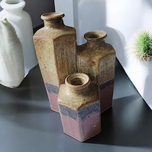 May include: A set of three ceramic vases with a rustic, textured appearance. The vases are rectangular with a light brown top, a dark blue band, and a reddish-pink base. The vases vary in size, with the tallest one in the center.