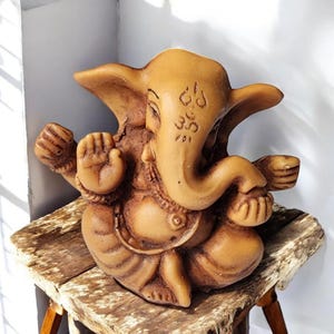 May include: A detailed, brown statue of the Hindu deity Ganesha, seated with multiple arms and an elephant head. The statue has intricate carvings and is displayed on a rustic wooden surface.