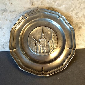 May include: A vintage pewter plate with an octagonal shape and a detailed embossed cityscape in the center. The plate features a raised rim and a central medallion depicting buildings and a clock tower. The overall color is a muted silver.