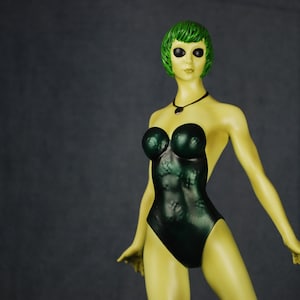 May include: A green-skinned figurine with green hair and large black eyes, wearing a dark green one-piece swimsuit. The figurine has a necklace with a dark green pendant.
