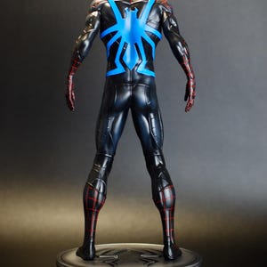 May include: A black and blue superhero figurine with red accents. The figure is posed from the back, showcasing a blue spider emblem on the back and red web-like detailing. The figurine is mounted on a silver circular base.