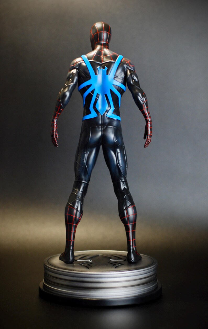 May include: A black and blue superhero figurine with red accents. The figure is posed from the back, showcasing a blue spider emblem on the back and red web-like detailing. The figurine is mounted on a silver circular base.