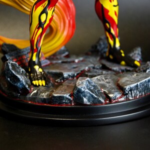 May include: A detailed figurine featuring legs and a flowing tail in vibrant yellow, red, and black hues. The figure stands on a rocky base with a red lava-like design, set on a black circular platform. The overall aesthetic suggests a fantasy or mythological theme.