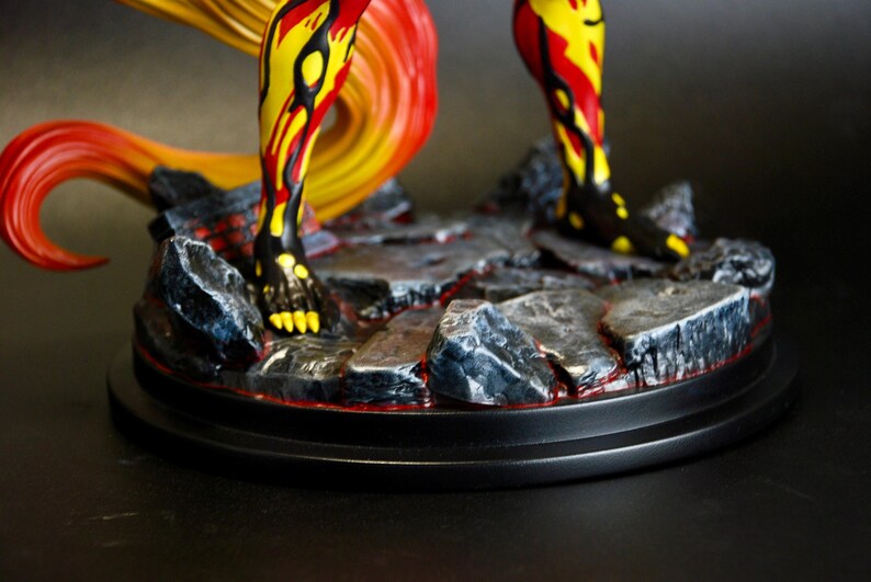 May include: A detailed figurine featuring legs and a flowing tail in vibrant yellow, red, and black hues. The figure stands on a rocky base with a red lava-like design, set on a black circular platform. The overall aesthetic suggests a fantasy or mythological theme.
