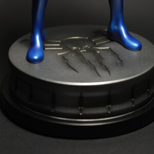 May include: A blue figurine standing on a black and silver base with a spider web design. The base has a raised spider web design with a skull in the center.