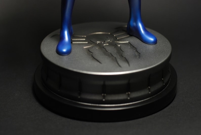 May include: A blue figurine standing on a black and silver base with a spider web design. The base has a raised spider web design with a skull in the center.