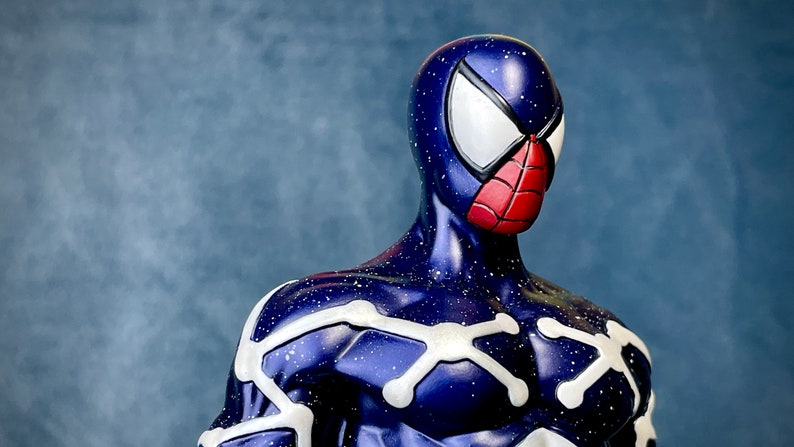Puede incluir: Una figura de superh&eacute;roe azul y blanca con un dise&ntilde;o de ara&ntilde;a roja en la m&aacute;scara. La figura tiene un patr&oacute;n de cielo estrellado en el traje.