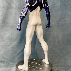 Puede incluir: Una figura de superh&eacute;roe blanca y azul con un patr&oacute;n estrellado. La figura est&aacute; de pie sobre una base plateada con un dise&ntilde;o circular.