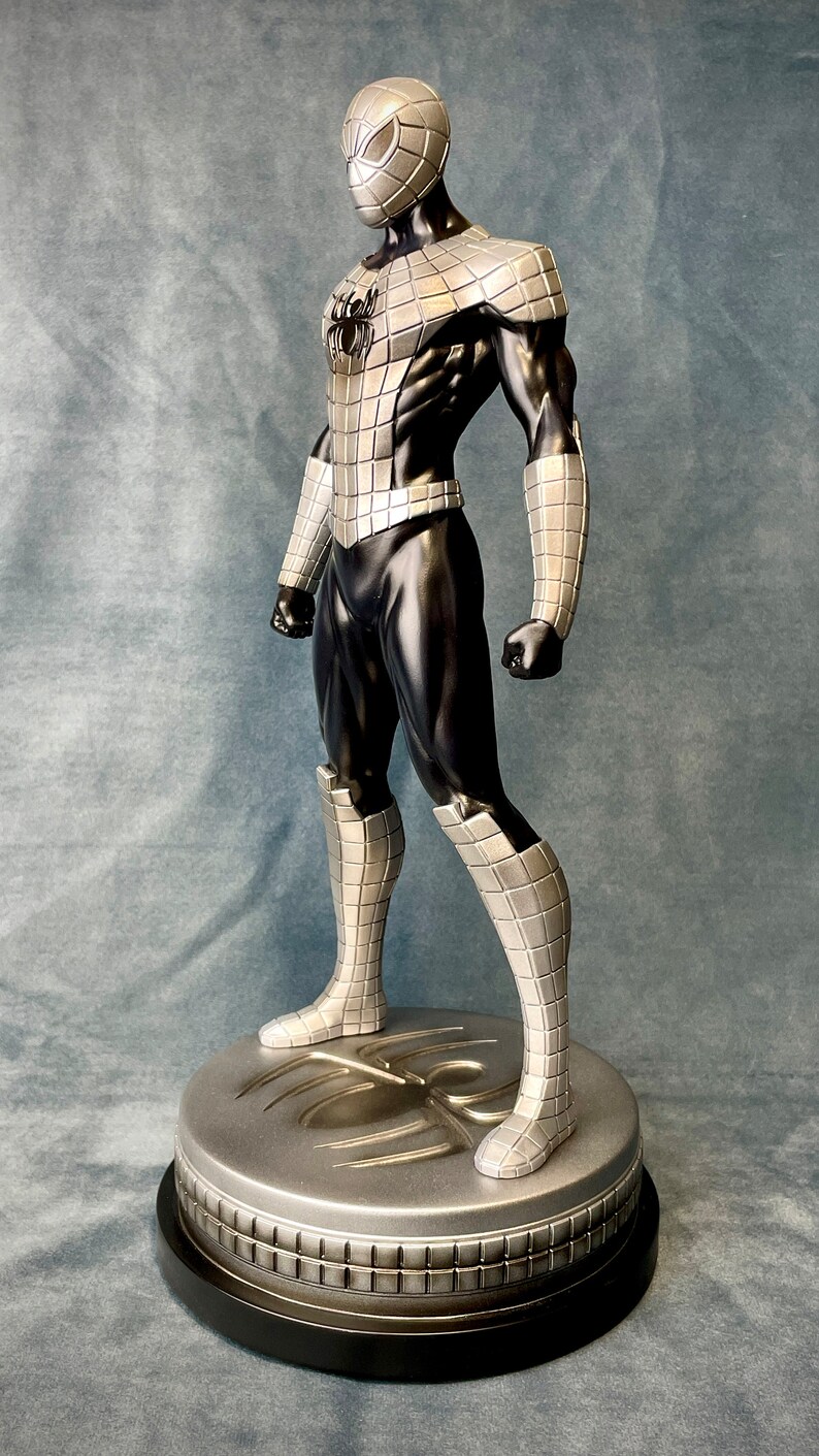 May include: A silver and black action figure of Spider-Man standing on a silver base with a spider web design. The figure is posed in a dynamic stance with one leg forward and arms raised.