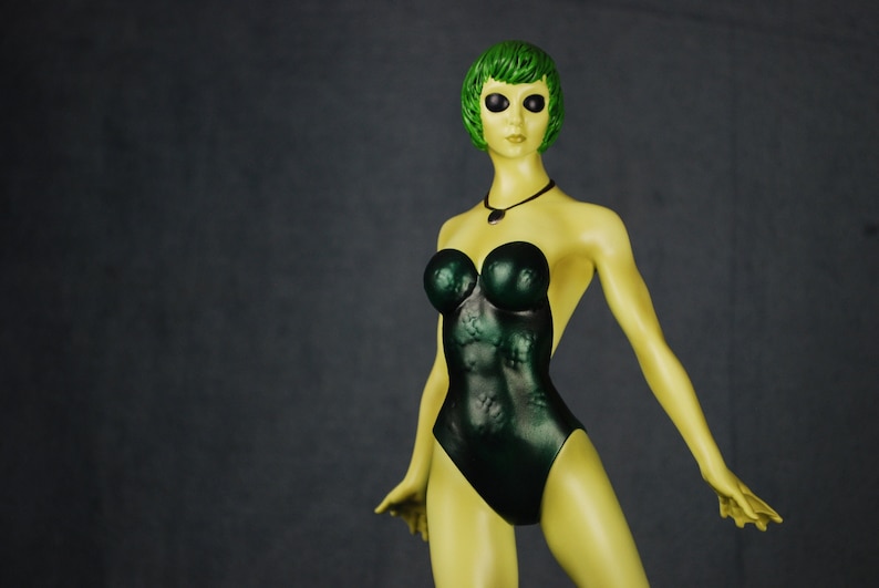 May include: A green-skinned figurine with green hair and large black eyes, wearing a dark green one-piece swimsuit. The figurine has a necklace with a dark green pendant.