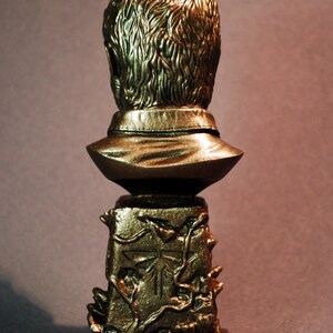 Pu&ograve; includere: Un busto dorato di un uomo con i capelli corti, che indossa una camicia con colletto. Il busto &egrave; montato su una base con un design dettagliato di viti e un simbolo di lucciola. La base &egrave; incisa con il testo "CUSTOM JOEL BUST".