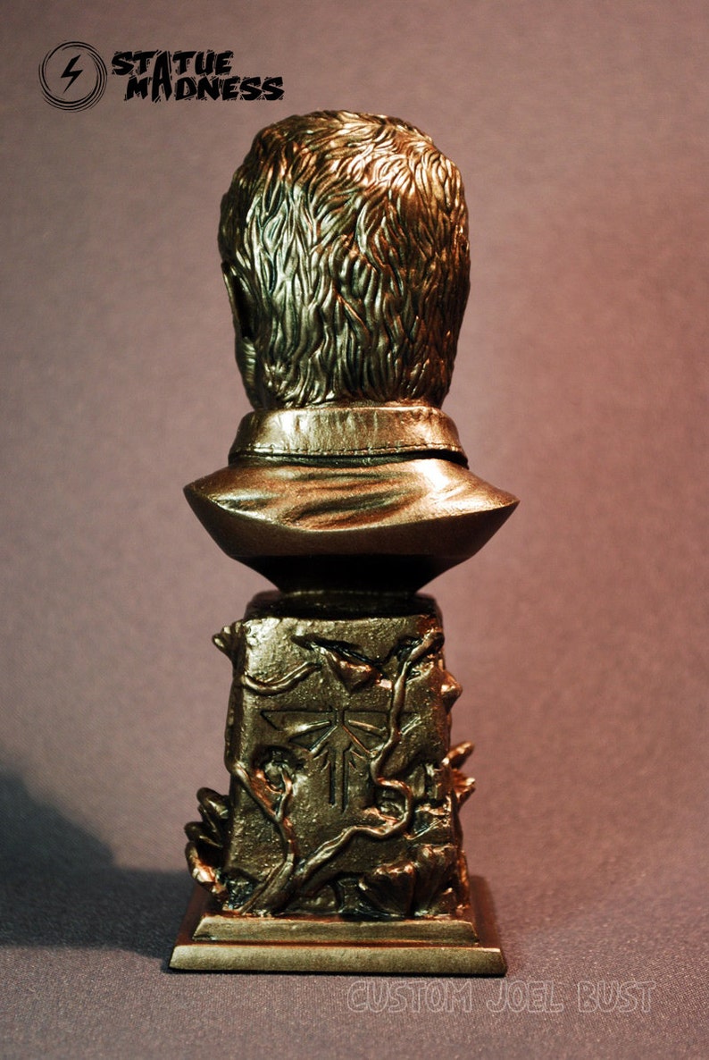 Pu&ograve; includere: Un busto dorato di un uomo con i capelli corti, che indossa una camicia con colletto. Il busto &egrave; montato su una base con un design dettagliato di viti e un simbolo di lucciola. La base &egrave; incisa con il testo "CUSTOM JOEL BUST".