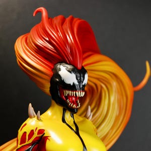 May include: A detailed figurine of a female character with a black and white face, red mouth, and spiky teeth. The figure has a yellow torso, red and yellow accents, and long, flowing hair in shades of red and yellow. The figure has sharp, spiky shoulder pads.